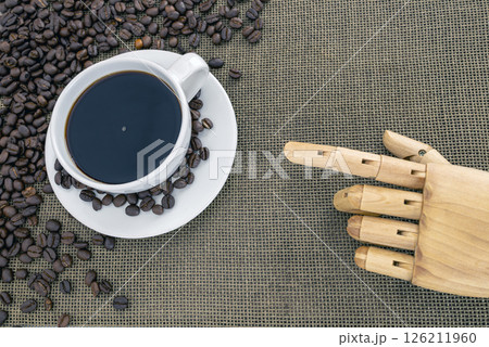 White coffee cup filled with black coffee sits on saucer surrounded by coffee beans on textured surface, with wooden hand pointing towards it 126211960