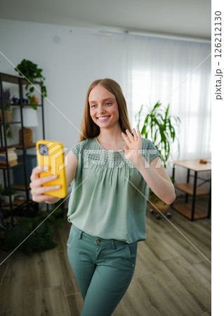 Smiling woman waves while taking a selfie in bright living room 126212130