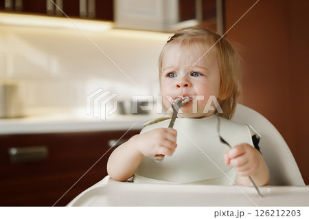 Infant girl or toddler sitting on high chair. Eating food by herself using spoon 126212203