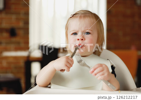 Infant girl or toddler sitting on high chair. Eating food by herself using spoon 126212217