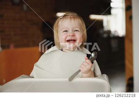 Infant girl or toddler sitting on high chair. Eating food by herself using spoon 126212218