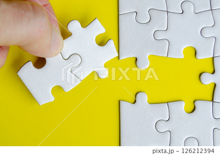 Hand matching white puzzle pieces lying on a vibrant yellow surface highlighting contrast and precise alignment. 126212394