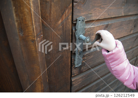 A little girl in pink jacket holding old metal door handle on the wood door in winter. Vintage iron pen. A little girl in pink jacket holding old metal door handle on the wood door in winter. Vintage iron pen. 126212549