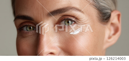 Closeup of middle aged woman with green eyes applying anti aging cream under her eye, showing natural skin texture and wrinkles 126214003