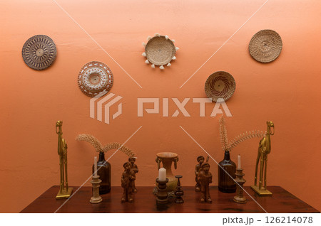 Interior wall decorated with woven baskets and various ornaments creating warm and artistic display with wooden figurines and brass sculptures on wooden table 126214078