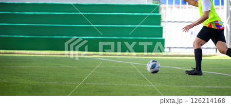 Soccer player running on green grass field with ball near feet during game action on outdoor stadium with green bleachers in background 126214168