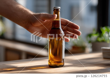 Hand reaches for beer bottle on sunlit wooden table, creating warm and inviting atmosphere. sunlight highlights amber color of beverage, adding cozy feel 126214755
