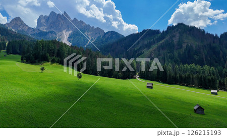 Majestic mountain landscape with lush green fields and rustic cabins under a bright blue sky in the Dolomites 126215193