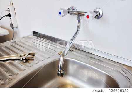 Newly installed dual handle kitchen tap with curved spout is fixed above stainless steel sink while adjustable wrench used during plumbing work remains on wet draining surface. 126216380