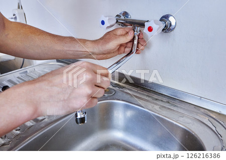 Male hands are tightening locking nut of faucet spout during installation of new dual handle kitchen tap above stainless steel sink with water traces on draining board. Male hands are tightening locking nut of faucet spout during installation of new dual handle kitchen tap above stainless steel sink with water traces on draining board. 126216386
