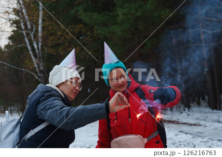 Two cheerful individuals share a playful moment in a snowy landscape, wearing festive hats while holding sparklers. Two cheerful individuals share a playful moment in a snowy landscape, wearing festive hats while holding sparklers. 126216763