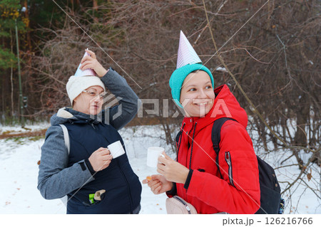 Friends savor the joy of winter in a snowy forest, wearing colorful party hats as they share warm drinks and snacks. Friends savor the joy of winter in a snowy forest, wearing colorful party hats as they share warm drinks and snacks. 126216766