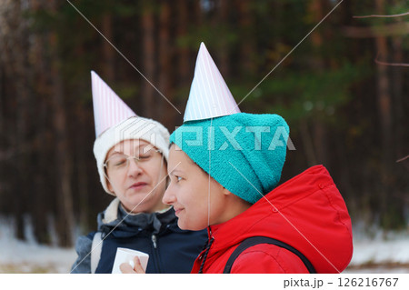 Friends savor the joy of winter in a snowy forest, wearing colorful party hats as they share warm drinks and snacks. 126216767
