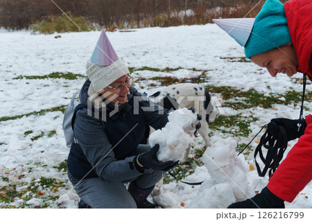 Friends gather in a snowy park, donning festive hats, as they joyfully build a snowman together. 126216799