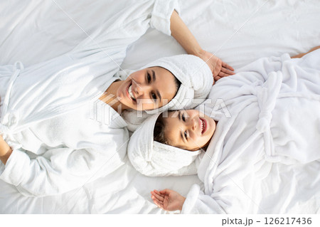 Happy young mother and little daughter in bathrobes and towels on heads laying on bed face to face and smiling at camera, top view. Cheerful mom and kid having beauty day at home 126217436