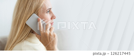 A woman with long blonde hair speaks on her smartphone while sitting comfortably in a bright and cozy room, copy space 126217445