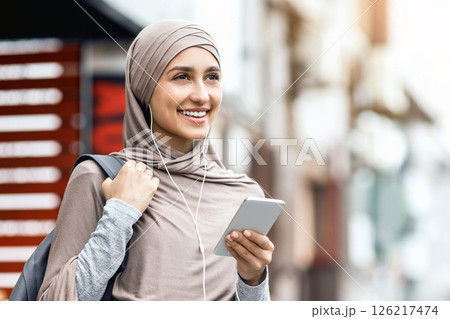 Positive arab girl in hijab using earphones and newest smartphone, walking by street and listening to music, empty space Positive arab girl in hijab using earphones and newest smartphone, walking by street and listening to music, empty space 126217474