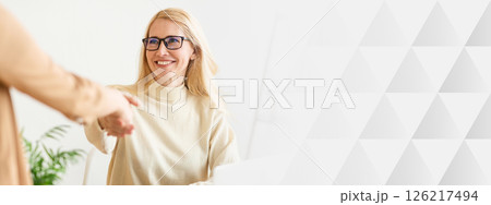 A woman with long blonde hair and glasses sits at a desk in a bright office. She is smiling warmly while extending her hand for a handshake, welcoming a visitor with friendliness, copy space 126217494