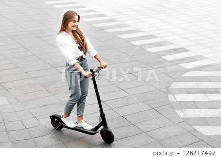 High angle view of cheerful casual girl riding black stand-up scooter on brick road, empty space for ad or text High angle view of cheerful casual girl riding black stand-up scooter on brick road, empty space for ad or text 126217497
