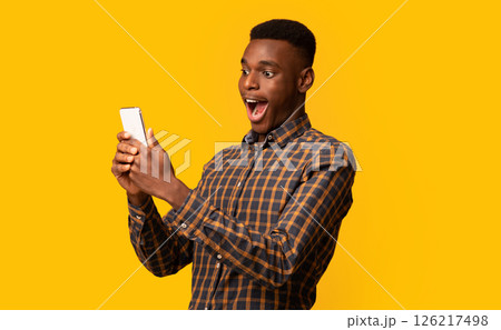 Mobile Offer. Surprised funny black guy looking at smartphone screen with excitement, emotionally reacting to online news, standing with opened mouth isolated over yellow background, copy space 126217498