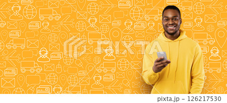 A cheerful buyer stands against a vibrant yellow background filled with delivery and shopping icons. He holds a smartphone, ready to complete an online purchase for a service. A cheerful buyer stands against a vibrant yellow background filled with delivery and shopping icons. He holds a smartphone, ready to complete an online purchase for a service. 126217530