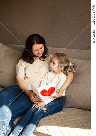 Young girl with light hair hugs mom tightly while holding up her childish drawing. Pure mother-daughter love. Young girl with light hair hugs mom tightly while holding up her childish drawing. Pure mother-daughter love. 126218746