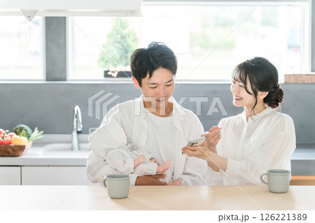 赤ん坊を抱っこする男性・パパとスマホの画面を見せて笑顔で会話する女性・ママ 赤ん坊を抱っこする男性・パパとスマホの画面を見せて笑顔で会話する女性・ママ 126221389