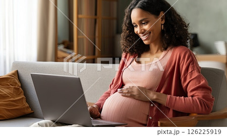 Happy pregnant black mom working remotely on laptop. Online video call. The smiling mom embodies the spirit of working mothers as she connects online during her pregnancy. Happy pregnant black mom working remotely on laptop. Online video call. The smiling mom embodies the spirit of working mothers as she connects online during her pregnancy. 126225919