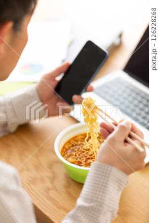 オフィスで仕事をしながらカップラーメンを食べるミドル男性 126228268