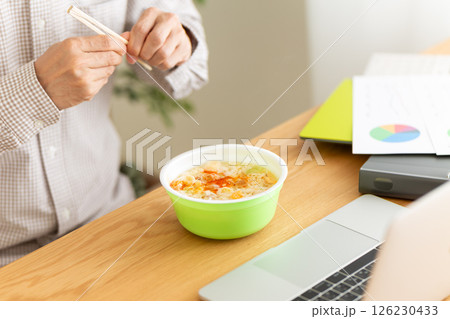 オフィスで仕事をしながらカップラーメンを食べるミドル男性 オフィスで仕事をしながらカップラーメンを食べるミドル男性 126230433