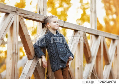 Little smiling caucasian child wearing jacket and trousers is posing at wooden bridge outdoors background. Little smiling caucasian child wearing jacket and trousers is posing at wooden bridge outdoors background. 126230978