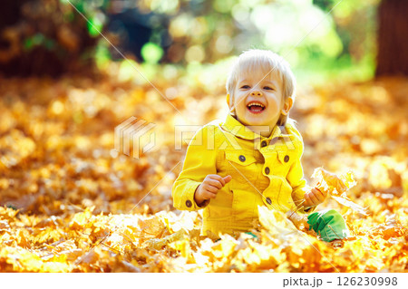 Little happy boy in yellow jacket is playing with leaves at golden autumn park background. Little happy boy in yellow jacket is playing with leaves at golden autumn park background. 126230998