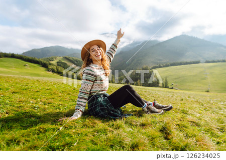 Stylish female traveler in hat has fun on top of hill, enjoys mountain landscape. Active lifestyle. 126234025
