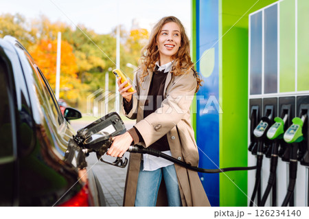 Beautiful woman filling car with gasoline at gas station. Online pay. Woman using app on phone. 126234140