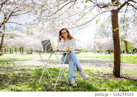 A young woman at a table in a blooming park works with a laptop. Freelancing, weekend concept. A young woman at a table in a blooming park works with a laptop. Freelancing, weekend concept. 126234320