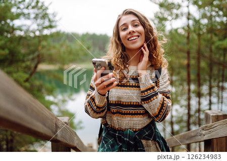 A young woman in a cozy sweater walking near a beautiful lake, smiling and using her phone 126234983