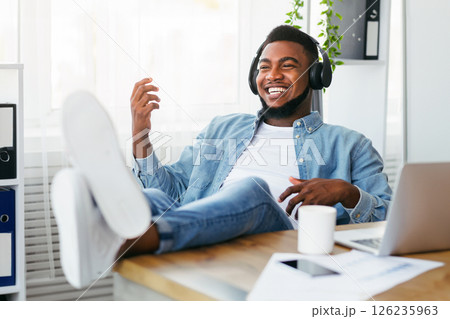 Break at work. Cheerful african american employee listening music in headphones and playing virtual guitar, relaxing at workplace 126235963