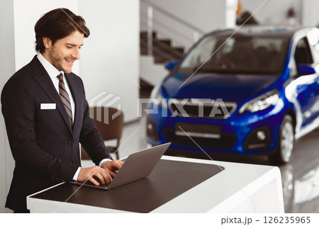 Salesman At Work. Car Dealer Standing At Desk In Luxury Dealership Office, Using Laptop, Empty space Salesman At Work. Car Dealer Standing At Desk In Luxury Dealership Office, Using Laptop, Empty space 126235965
