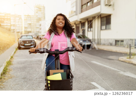 A cheerful lady with curly hair enjoys riding her bicycle along a city street. The sun shines brightly as she navigates past parked cars and buildings, showcasing her playful spirit. A cheerful lady with curly hair enjoys riding her bicycle along a city street. The sun shines brightly as she navigates past parked cars and buildings, showcasing her playful spirit. 126236017