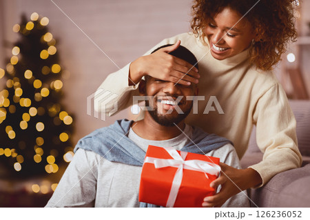 Xmas surprise. Loving wife giving gift to husband and closing his eyes in front of christmas tree Xmas surprise. Loving wife giving gift to husband and closing his eyes in front of christmas tree 126236052