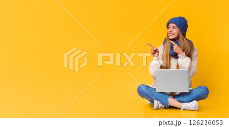 Beautiful young woman in knitted hat with laptop on her lap pointing at empty space, pink background 126236053