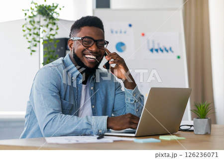 Happy Black Entrepreneur Speaking On Mobile Phone While Working On Laptop In Office. Happy Black Entrepreneur Speaking On Mobile Phone While Working On Laptop In Office. 126236071