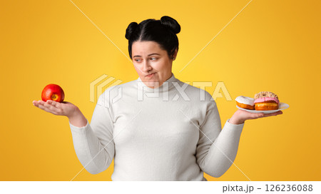 Complex Choice. Hungry Obese Girl On Diet Making Choice Between Donuts And Apple, Standing Over Yellow Background In Studio, Free Space 126236088