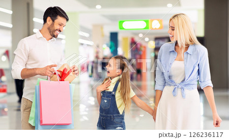 A father holding shopping bags and a gift with a red bow reveals it to his excited daughter, as his wife walks beside them. The three are shopping in a modern mall A father holding shopping bags and a gift with a red bow reveals it to his excited daughter, as his wife walks beside them. The three are shopping in a modern mall 126236117