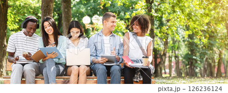 Multiethnic friends studying for exams together outdoors, sitting on bench in campus, having fun together 126236124