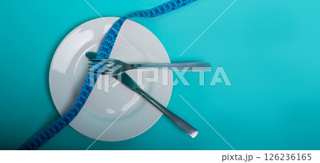 Healthy eating, weight loss obesity. An empty plate with a measuring tape in centimeters, a fork and a knife on a blue background from above. Diet menu.Place for an inscription. Advertising. Healthy eating, weight loss obesity. An empty plate with a measuring tape in centimeters, a fork and a knife on a blue background from above. Diet menu.Place for an inscription. Advertising. 126236165