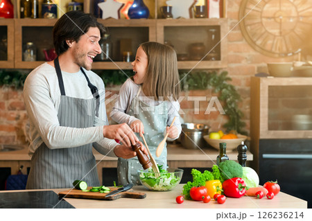 Happy father and little daughter cooking vegetable salad in kitchen, adding seasoning and laughing together while preparing lunch at home Happy father and little daughter cooking vegetable salad in kitchen, adding seasoning and laughing together while preparing lunch at home 126236214