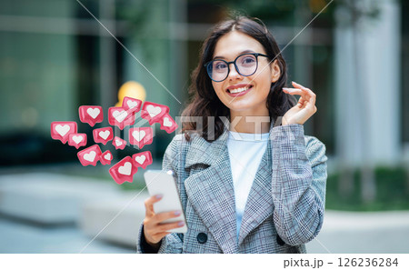 This image shows a woman in a grey blazer, holding a smartphone in her hand. She is smiling and looking at the device. There are several heart-shaped icons floating above her phone 126236284