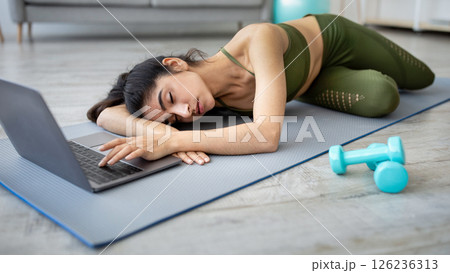 Tired young Indian woman sleeping on yoga mat in front of laptop, too exhausted from online training at home. Overworked Eastern lady taking break from exercising indoors 126236313
