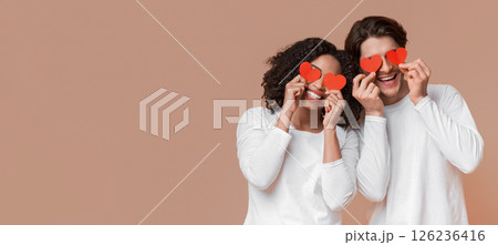 Blinded by love. Happy romantic interracial couple holding red hearts over eyes and smiling, posing together over yellow background with free space Blinded by love. Happy romantic interracial couple holding red hearts over eyes and smiling, posing together over yellow background with free space 126236416